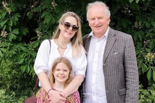 Philip standing outside in front of some green foliage with his daughter and granddaughter.