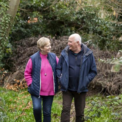 An older couple smiling and looking at each other as they walk arm in arm through woodland.