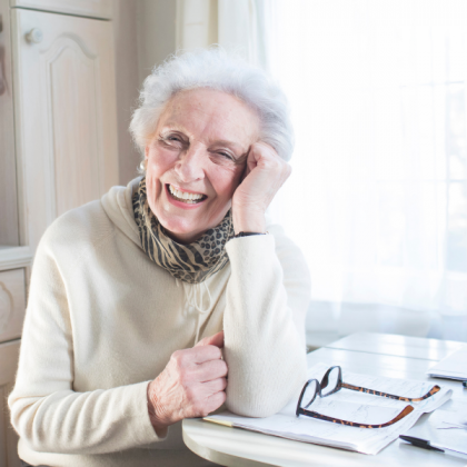 Photo of an older lady smiling with her elbow resting on a table with glasses and a book on it. 