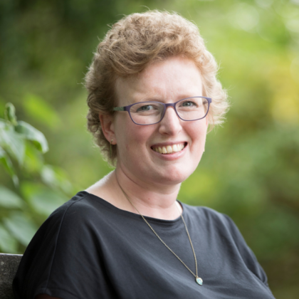 Headshot of Lizzie, smiling. She is wearing a grey top, a long necklace, square frame glasses and has short curly hair