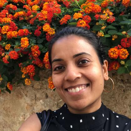 headshot of Melissa, smiling and standing in front of lots of orange flowers. She is wearing a black top and gold hoop earrings. 