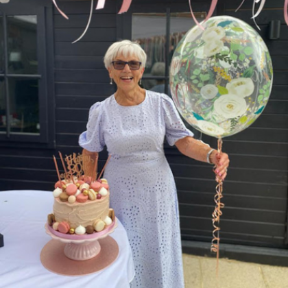 Jacky wearing a white dress posing with an 80th birthday cake and floral helium balloon