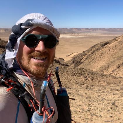 Selfie of Fred in the desert. He is has a ginger beard and has a big smile. He is wearing running goggles, a backpack with water bottle attachments and a white cloth headcover.