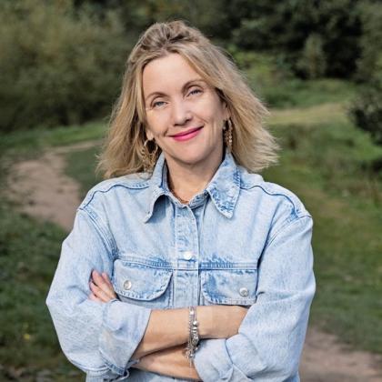 A woman in a stylish denim jacket, with short blonde hair smiling with arms folded