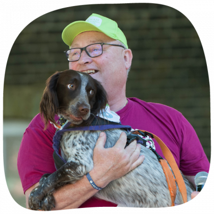 A Man wearing a banham marsden march cap and pink shirt smiling. He has glasses and is holding a brown and white collie dog