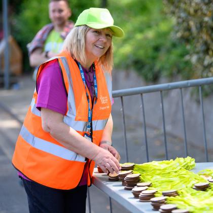 A woman smiling. She is wearing a Banham Marsden March volunteer vest and cap and by a table of finishers medals. 