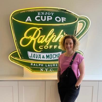 Fran smiling and standing in front of a Ralph Lauren coffee sign mounted to the wall, she's wearing a pink t-shirt with dungarees.