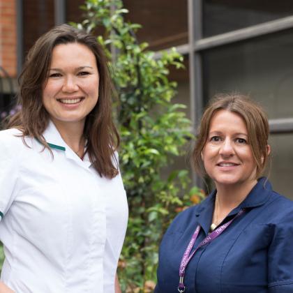 Two women stood outside smiling. The woman on the left is tall and wearing a white uniform. The woman on the left is shorter and wearing a navy blue nurses uniform.