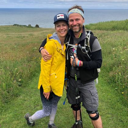 A young man and woman arm in arm. They are on a grassy hill with the sea in the background. They are wearing hiking gear, Royal Marsden Cancer Charity T-shirts and big smiles! Jackie is on the left and is also wearing a bright yellow coat. 
