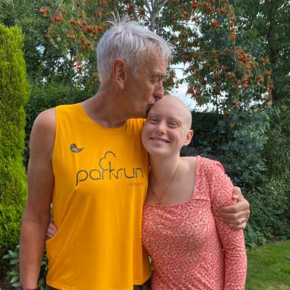 Chris with his daughter Alex. They are standing in a garden. Alex is smiling at the camera. Chris has his arm around Alex and is kissing Alex's head.