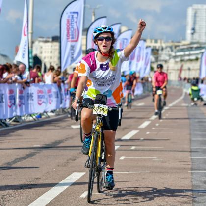 London to Brighton cycle ride cyclist cheers going through the finish line. She is wearing a blue helmet and a Royal Marsden Cycle vest.