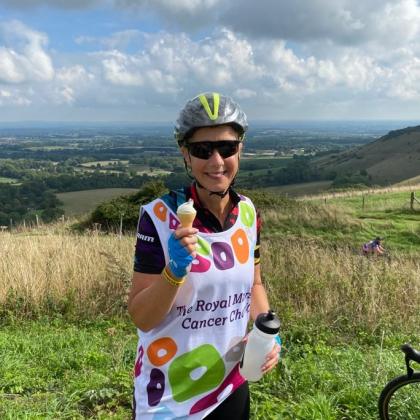 Carolyn enjoying a water break at the top of Ditchling Beacon. She is smiling and wearing her Royal Marsden cycling jersey, sunglasses and her cycling helmet. 