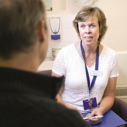 Clare sitting in a hospital consultation room holding a pen and folder, talking to a patient