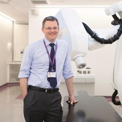 Professor Nicholas van As smartly dressed wearing a shirt and tie standing next to a CyberKnife machine - a large white robotic arm
