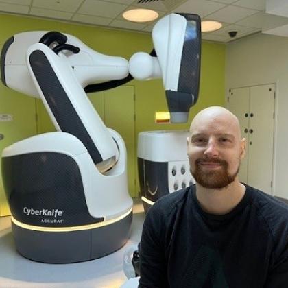 Alastair smiling and standing in front of a large white machine that resembles a robotic arm. 
