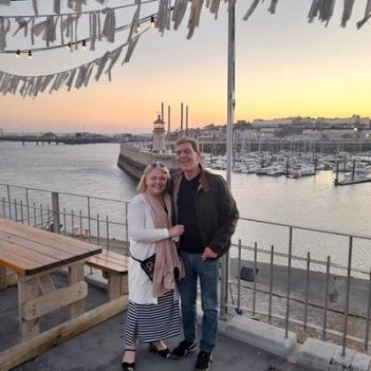 Keith and his wife on holiday, in front a view of a dock with moored boats at sunset