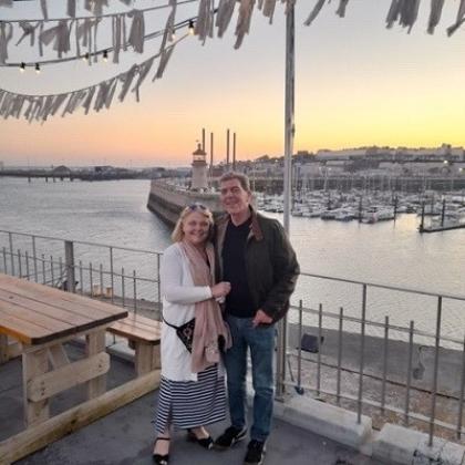 Keith and his wife on holiday, in front a view of a dock with moored boats at sunset 