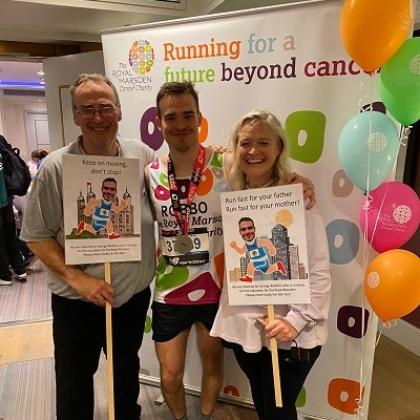 George standing in the middle of his parents, wearing his running gear and a marathon medal around his neck. They are in front of a Royal Marsden Cancer Charity roll-up banner and balloons and his parents are holding marathon support signs