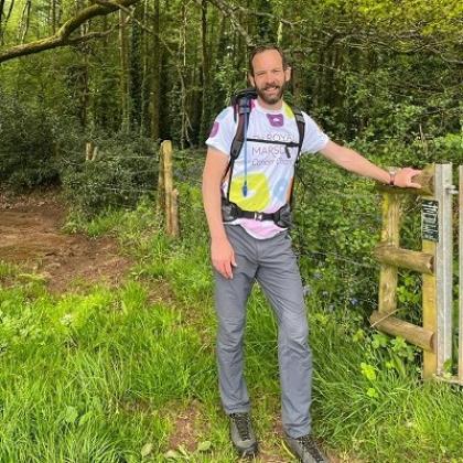 Jon leaning on a fence in a wooded area, wearing a backpack, walking gear and a Royal Marsden Cancer Charity t-shirt