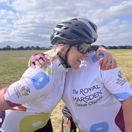 Two people wearing Royal Marsden Cancer Charity biking gear and helmets hugging each other and smiling