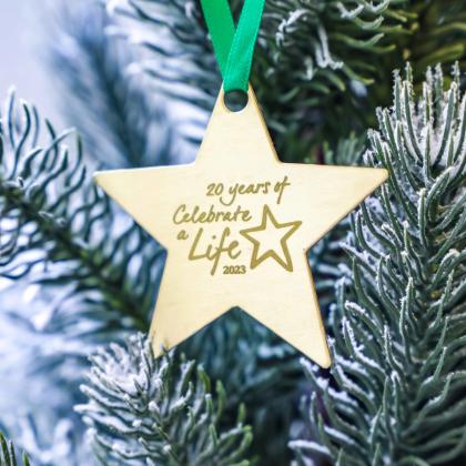 A brass star decoration on a Christmas tree. It has a green ribbon and '20 years of Celebrate a Life 2023' engraved on it.