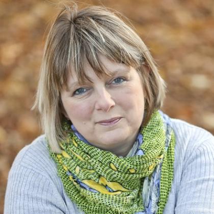 Head and shoulders image of a woman with short blonde hair, a grey jumper and green scarf.