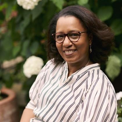 A Royal Marsden patient smiling and sitting in front of some potted flowers, wearing a stripped shirt and glasses with shoulder-length styled hair