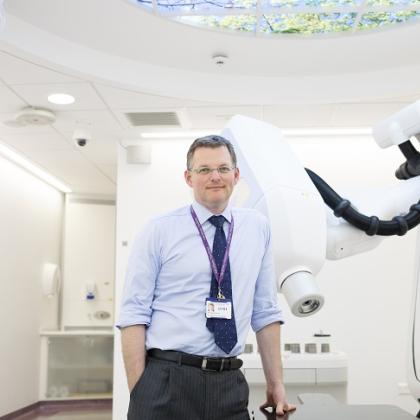A smartly-dressed Royal Marsden doctor with a lanyard, standing next to a large white machine with a robotic arm