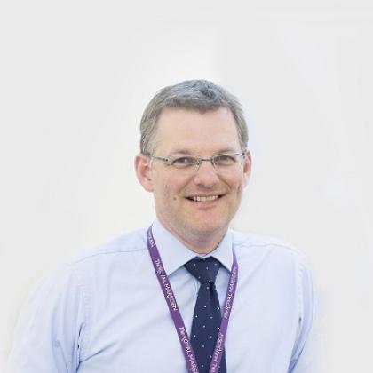 Headshot of a Royal Marsden doctor in a shirt and tie, wearing a purple lanyard 