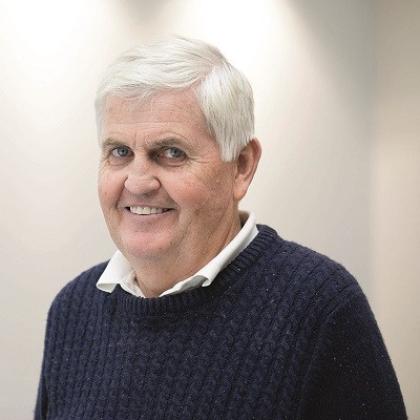 A smiling older man with grey hair and wearing a blue knitted jumper