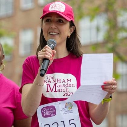 A person in a bright pink Banham Marsden March t-shirt and hat, holding a piece of paper and speaking to a crowd through a microphone