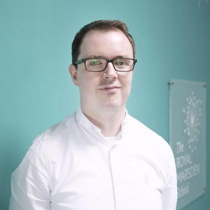 Headshot of a man in a white shirt and glasses, with a sign behind him that says 'The Royal Marsden School'