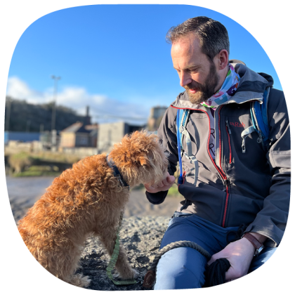 A man sitting on a beach, wearing a winter jacket and stroking a small fluffy ginger dog