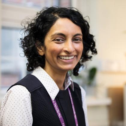 Portrait of a smiling woman, she has short dark curly hair and is wearing a purple Royal Marsden Hospital lanyard over her shirt and jumper. 