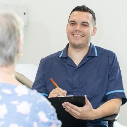 A man in a blue nurse uniform holding a clip board and pen, smiling and talking with a patient