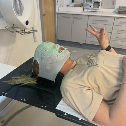 a person lying on a table in a hospital room, with a mask over her face and head. She's smiling and giving a peace sign