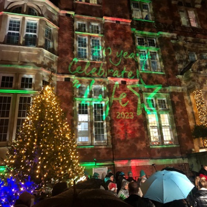 People gathered outside the front of The Royal Marsden in Chelsea with Christmas lights projected onto it saying '20 years Celebrate a Life 2023'. 