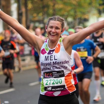 A person i a Royal Marsden Cancer Charity running shirt smiling and holding their hands up while running along a road with other marathon runners