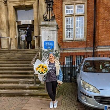A patient standing outside The Royal Marsden Chelsea front steps, smiling and holding a bunch of flowers