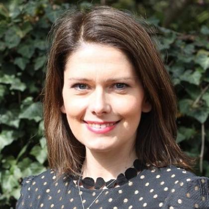 Headshot of Aneta smiling, she has shoulder-length brown hair and is wearing a black spotty top. She's in front of a green bush