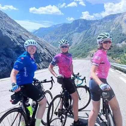 Three smiling people on bikes wearing cycling gear and helmets, stationary on a winding road surrounded by mountains