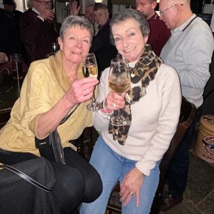 Two people sitting down in a busy pub, smiling and holding up glasses of wine