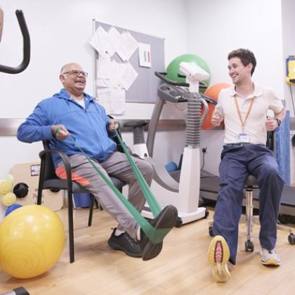 Naz with Adrian Fautly, both seated and smiling and surrounded by exercise equipment. Adrian is demonstrating how to use an exercise band.