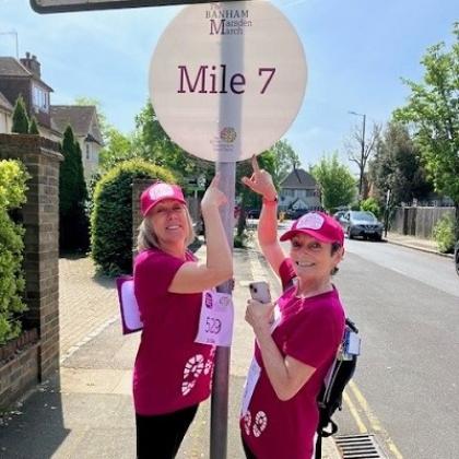 Two people in Banham Marsden march t-shirts and hats pointing to a sign that says 'mile 7' on a sunny day down a residential road in London