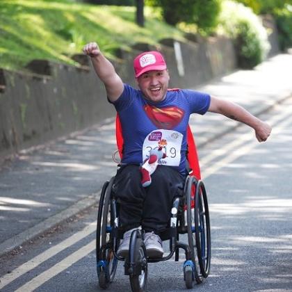 A Royal Marsden patient in a wheelchair, wearing a Banham Marsden March cap and dressed as superman, doing a superman pose with his fist outstretched