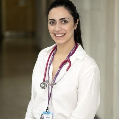 A Paediatric Registrar smiling and standing in a hospital corridor, wearing a smart shirt with an ID badge and stethoscope around her neck