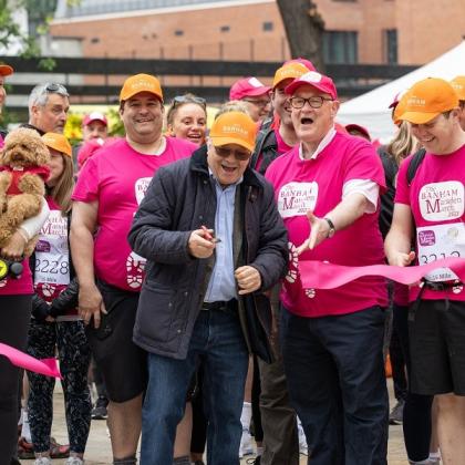 A group of people in Banham Marsden March outfits and hats with a man in front smiling and cutting a pink ribbon.