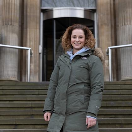 A smiling woman standing outside The Royal Marsden Hospital entrance. She has short brown curly hair and a khaki green coat on.