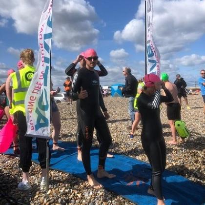 A man in a wetsuit, swimming cap and goggles on a pebbled beach giving a thumbs-up