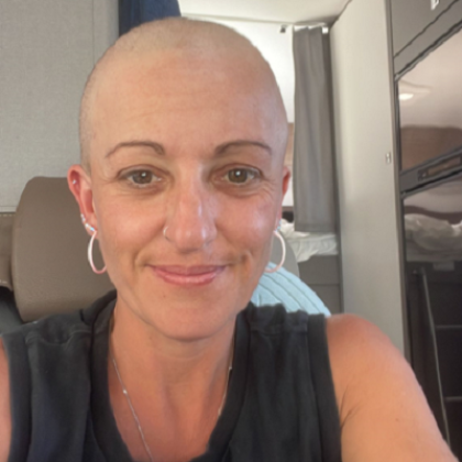 Head and shoulder of a woman smiling and sitting in a campervan, she's wearing a summery top and has no hair